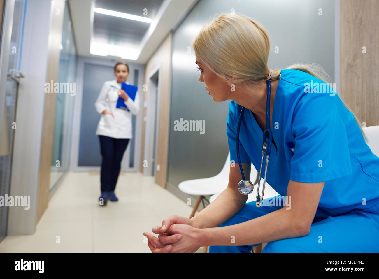 Surgeon sitting in hospital hallway, leaning forward, doctor walking along hallway Stock Photo