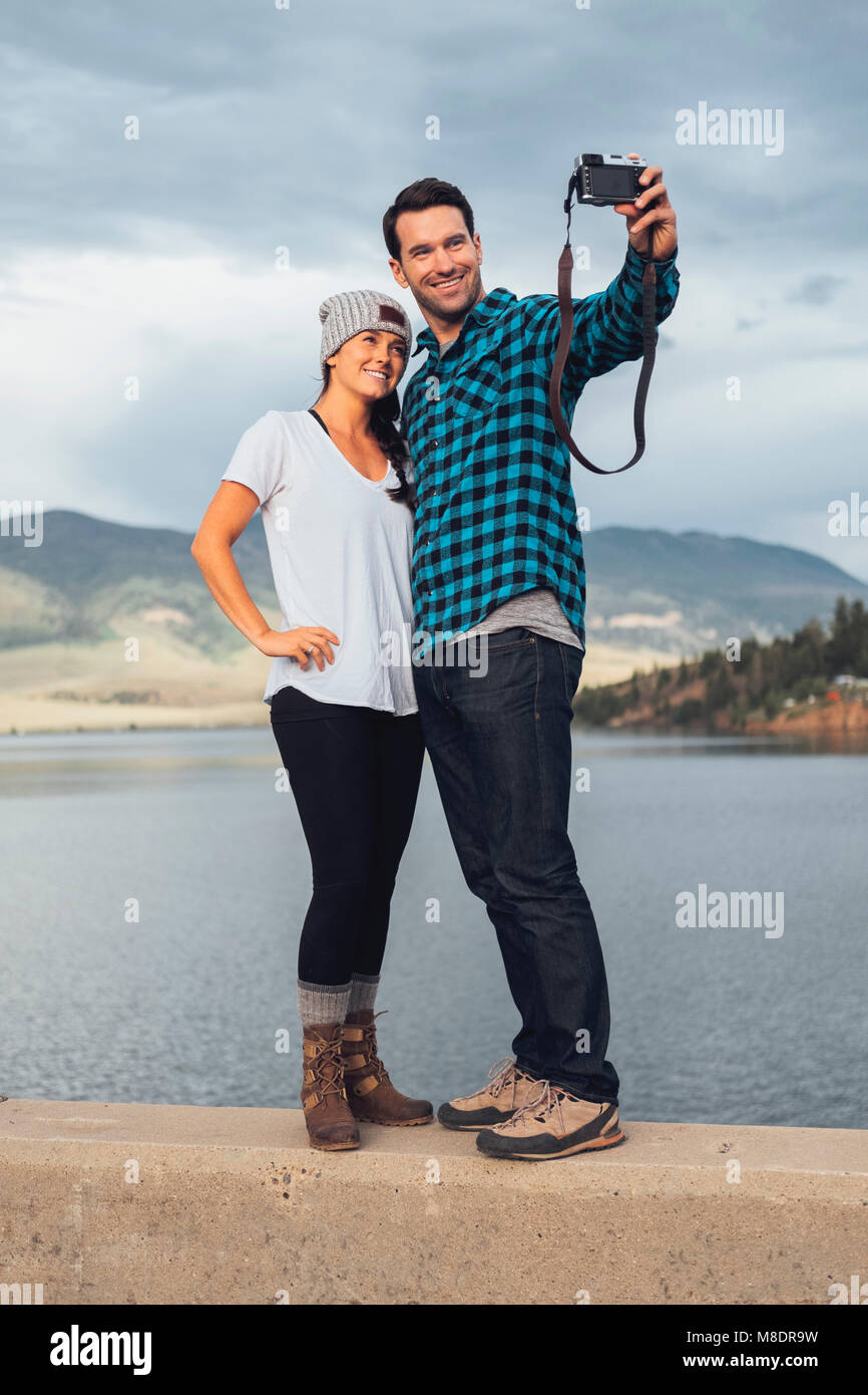 Couple standing on wall beside Dillon Reservoir, taking selfie, using camera, Silverthorne, Colorado, USA Stock Photo