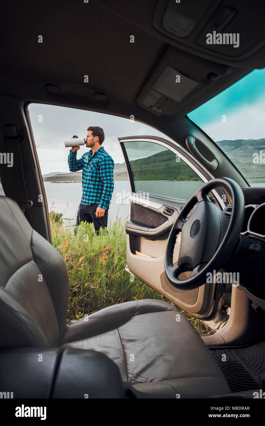 Mid adult man standing beside Dillon Reservoir, drinking from water bottle, view through parked car, Silverthorne, Colorado, USA Stock Photo