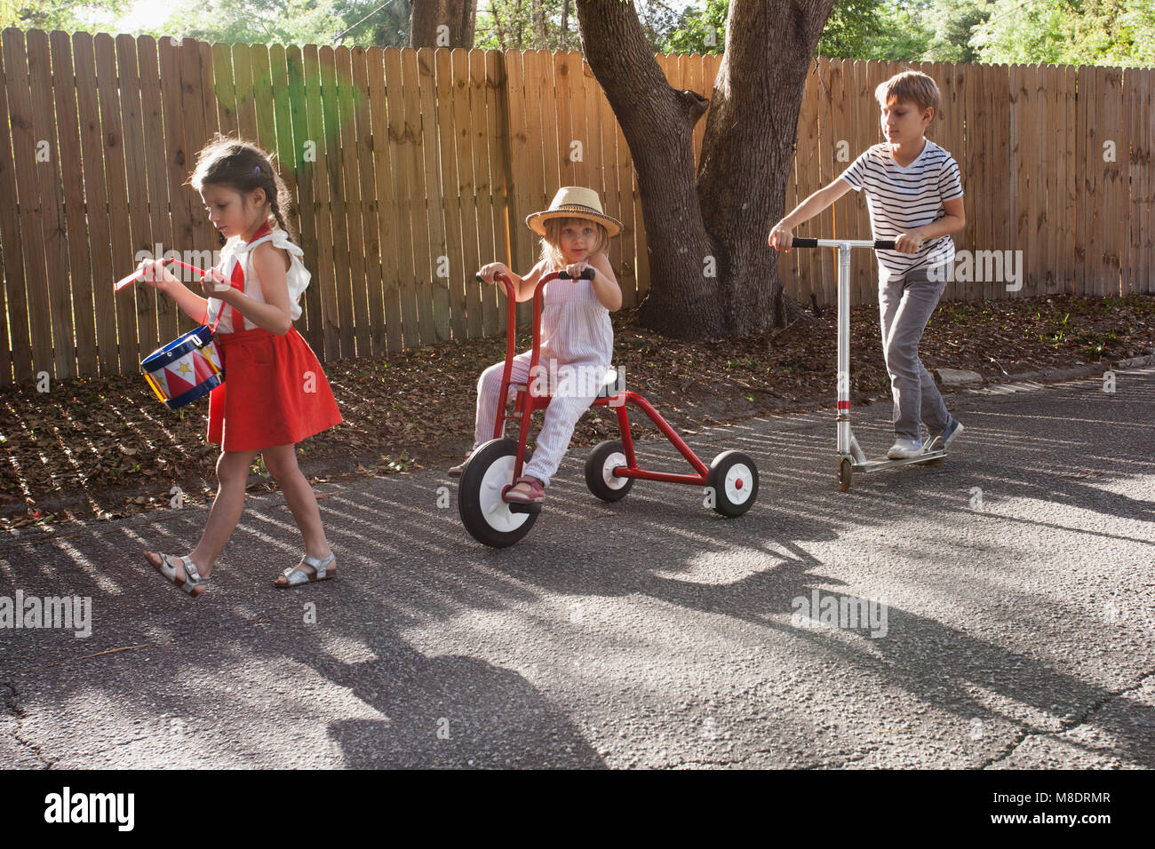 Three children in mini parade, banging drum, riding tricycle and using scooter Stock Photo