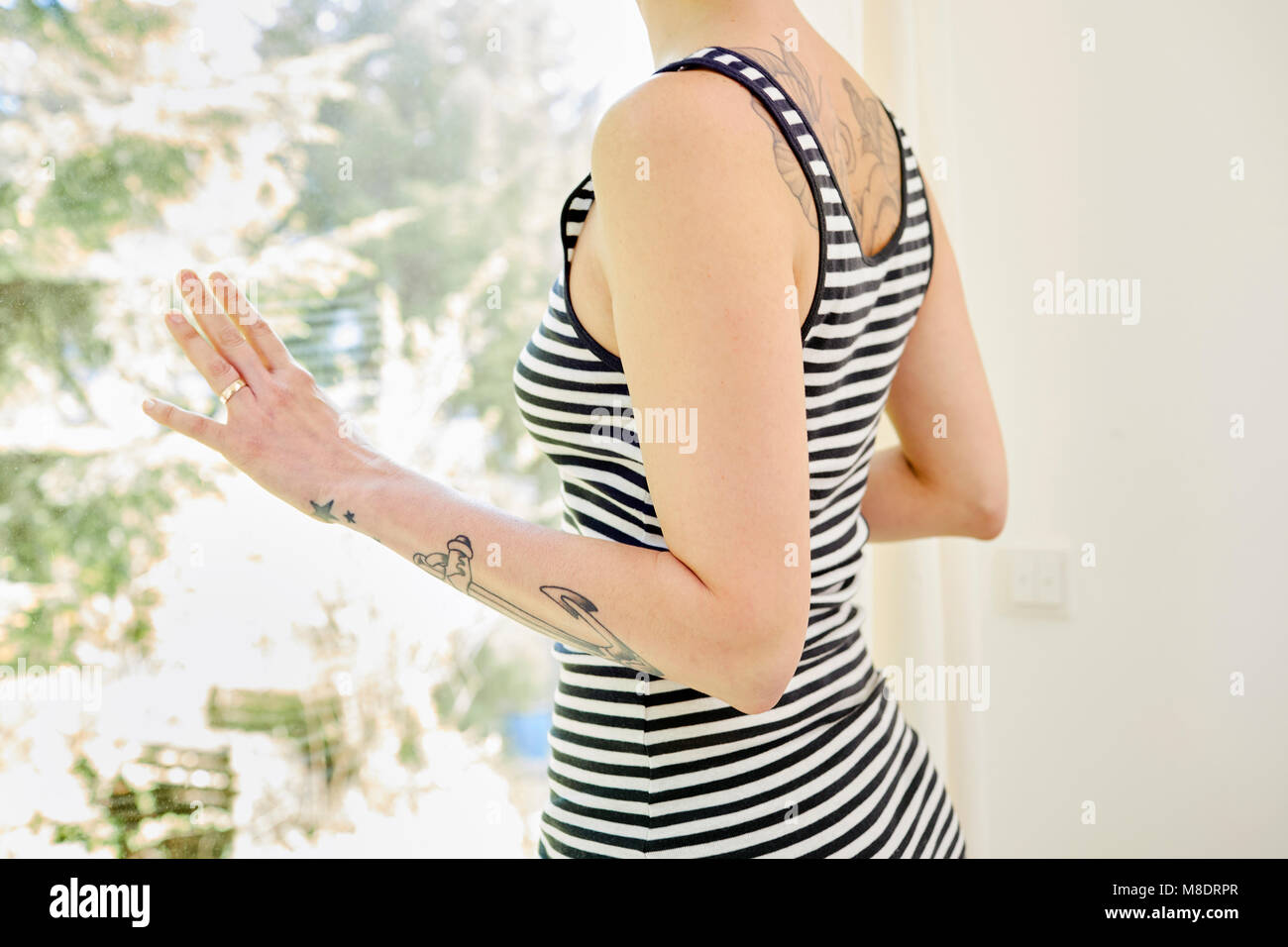 Mid adult woman, with tattoos on arm, looking out of window, mid section Stock Photo