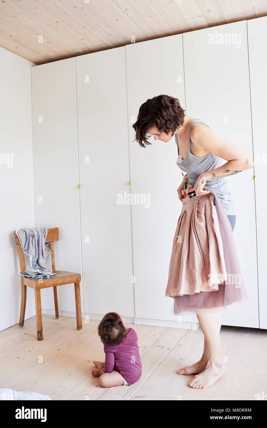 Mother holding skirt up against her, baby girl sitting on floor watching her Stock Photo