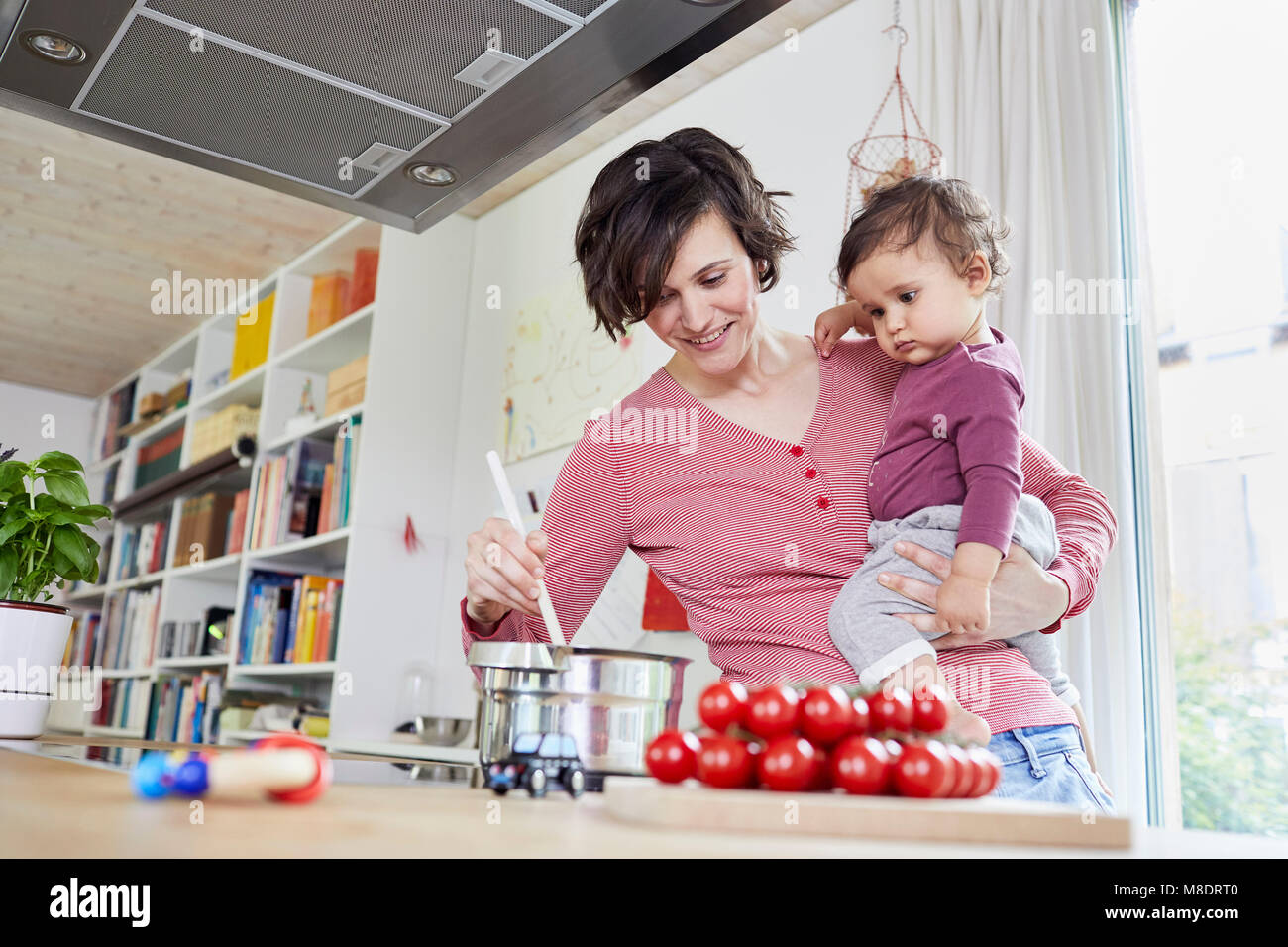 Mother holding baby girl in kitchen, whilst stirring pot on stove Stock Photo