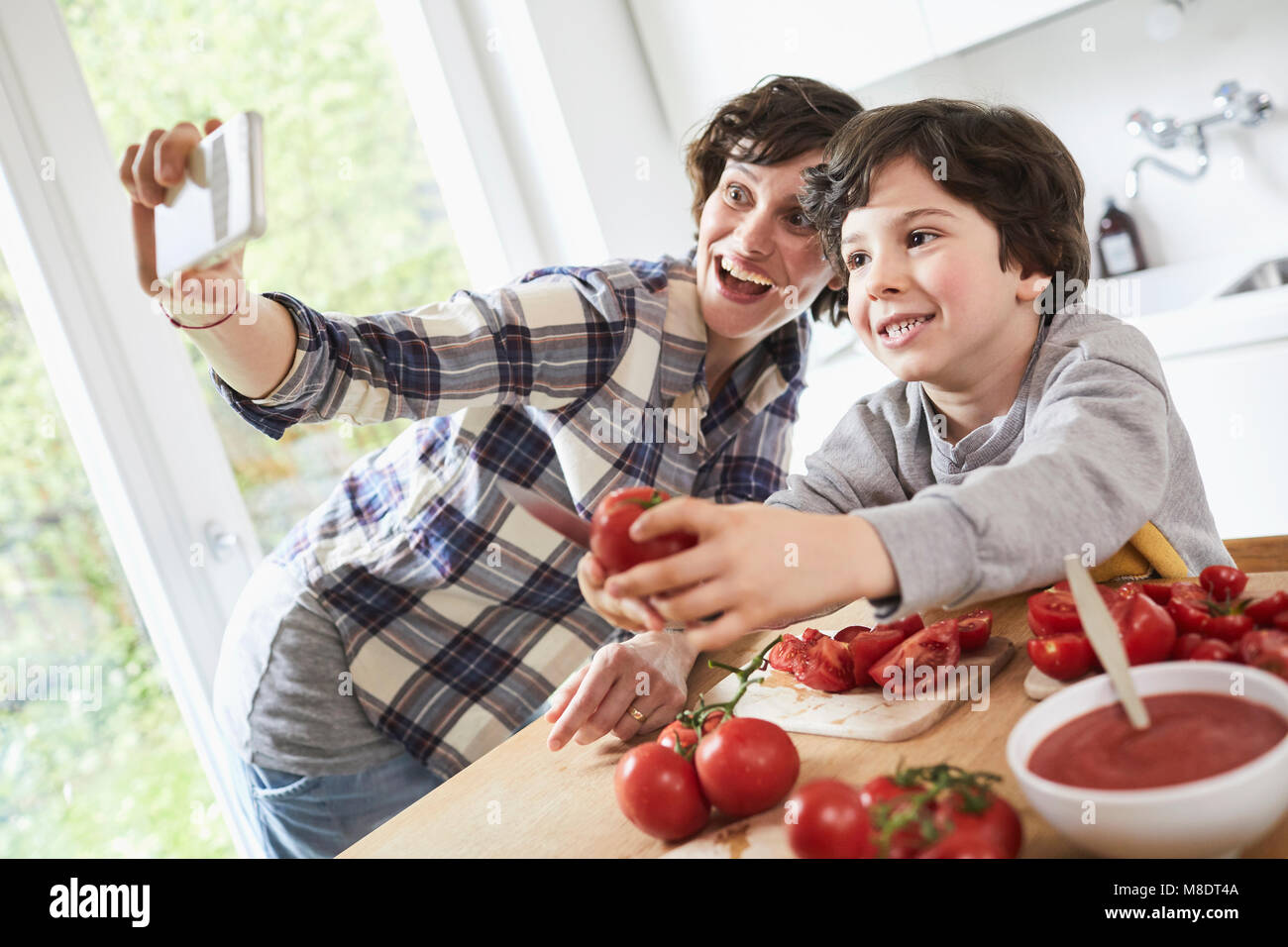 Mother and son preparing food in kitchen, mother taking selfie using smartphone Stock Photo
