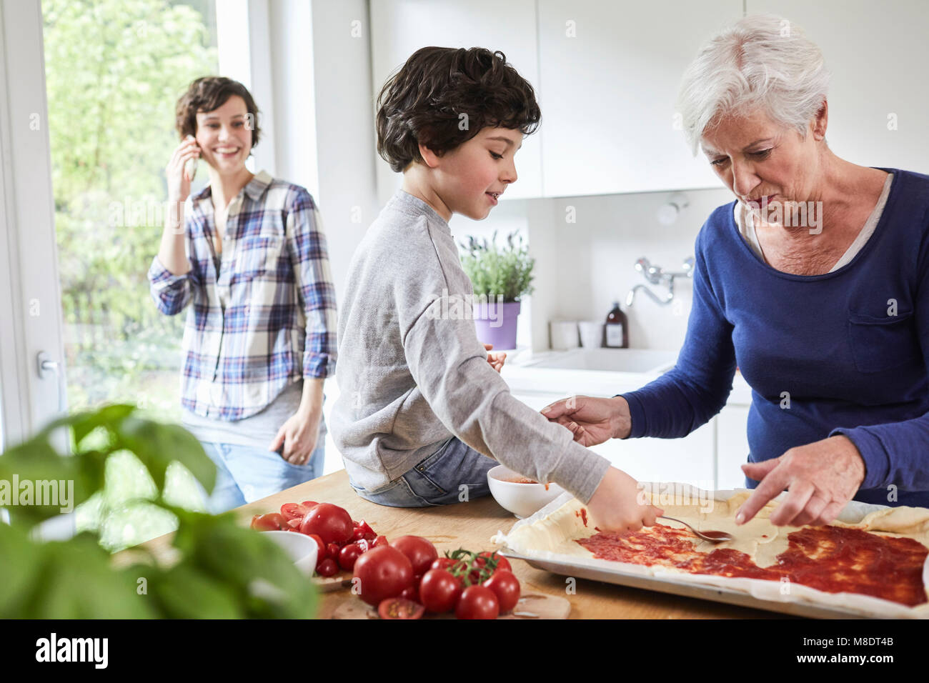 Grandmother and grandson making pizza in kitchen, mother in background using smartphone Stock Photo