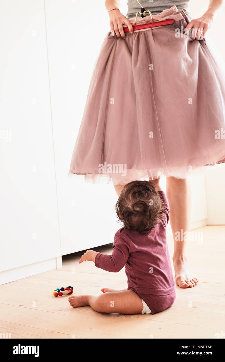 Mother holding skirt up against her, baby girl sitting on floor watching her, low section Stock Photo