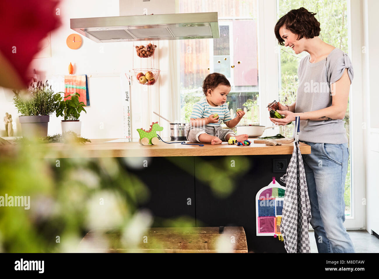 Baby daughter sitting on kitchen counter watching mother prepare vegetables Stock Photo