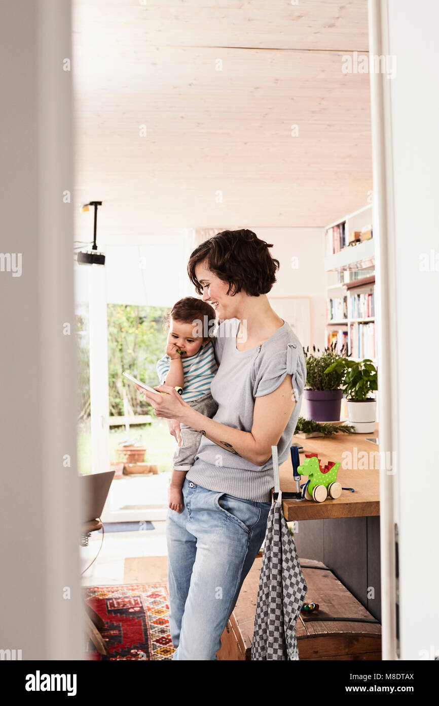 Mother holding baby daughter in kitchen, looking at smartphone Stock Photo