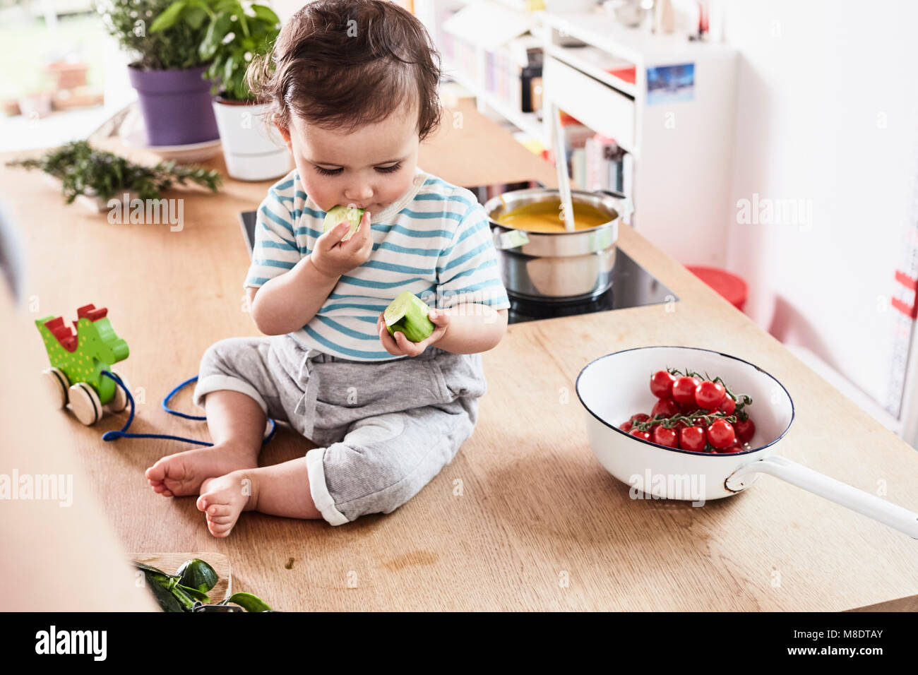 Baby girl sitting on kitchen counter, eating cucumber Stock Photo