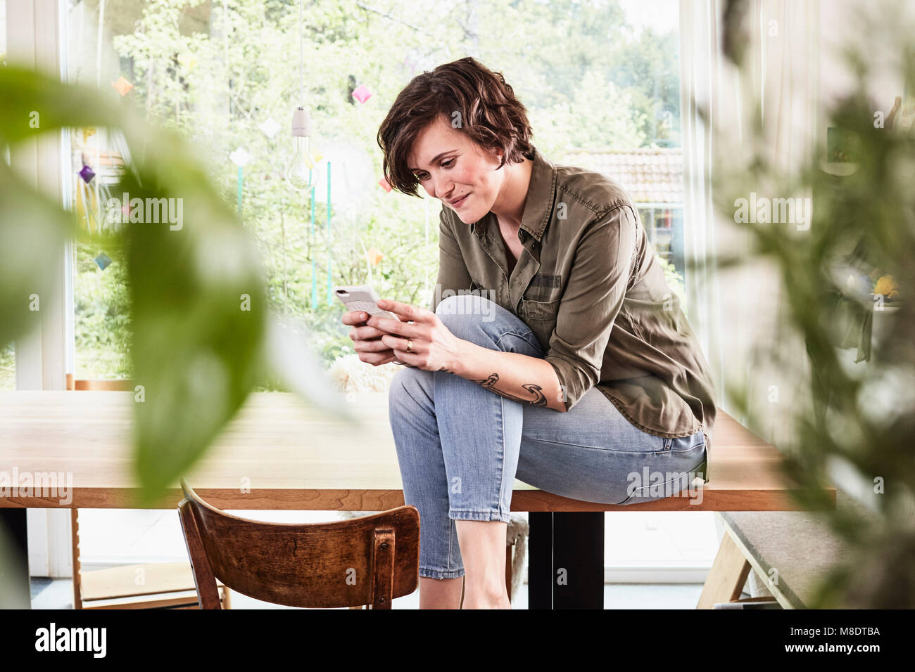 Mid adult woman at home, sitting on table, using smartphone Stock Photo