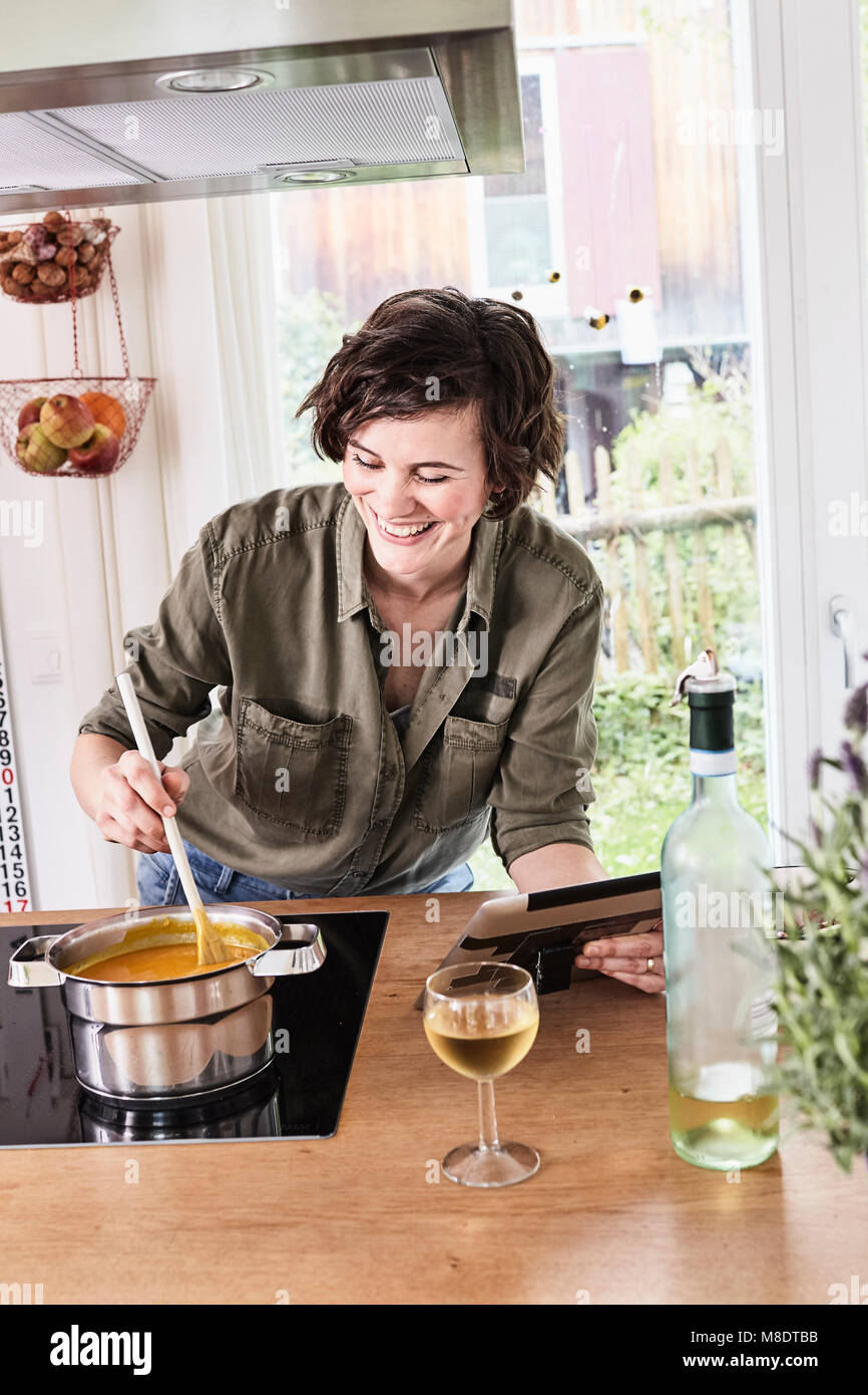 Mid adult woman stirring pot on stove in kitchen, holding digital tablet, laughing Stock Photo