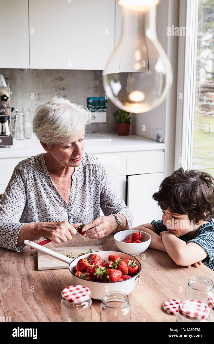 Grandmother sitting at kitchen table, preparing strawberries, grandson sitting beside her, watching Stock Photo