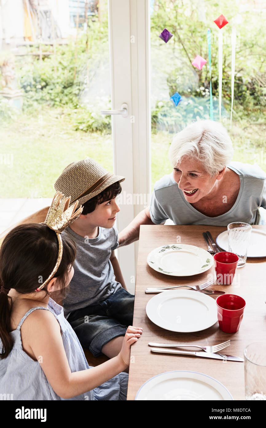 Grandmother sitting at kitchen table with grandson and granddaughter, smiling Stock Photo