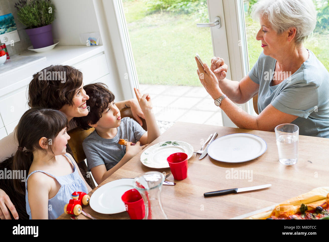 Grandmother sitting at kitchen, photographing grown daughter and grandchildren, using smartphone Stock Photo