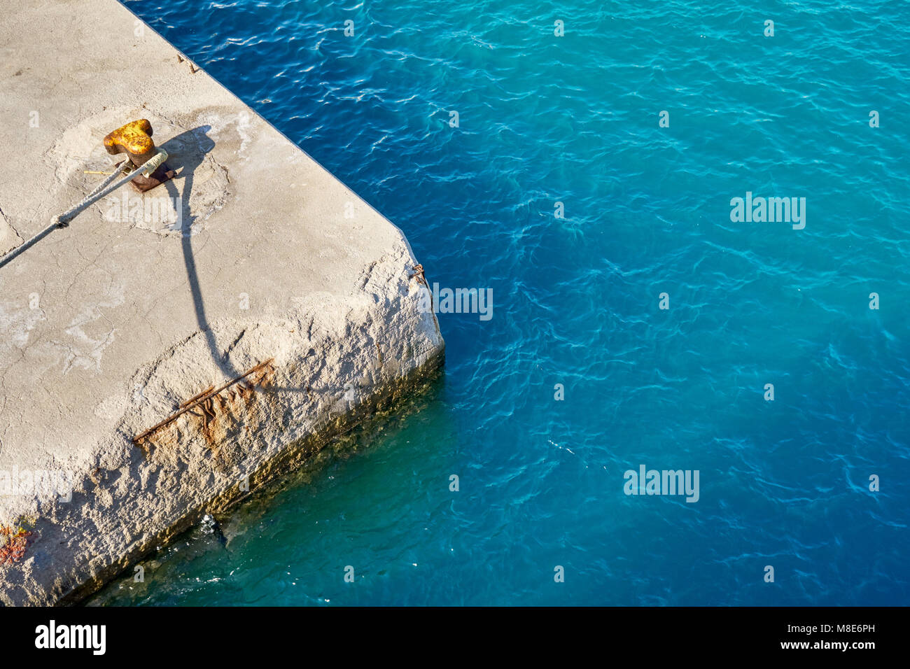 Yellow mooring bitt with rope on grey concrete pier at blue sea water with small waves under bright sunlight on summer day Stock Photo