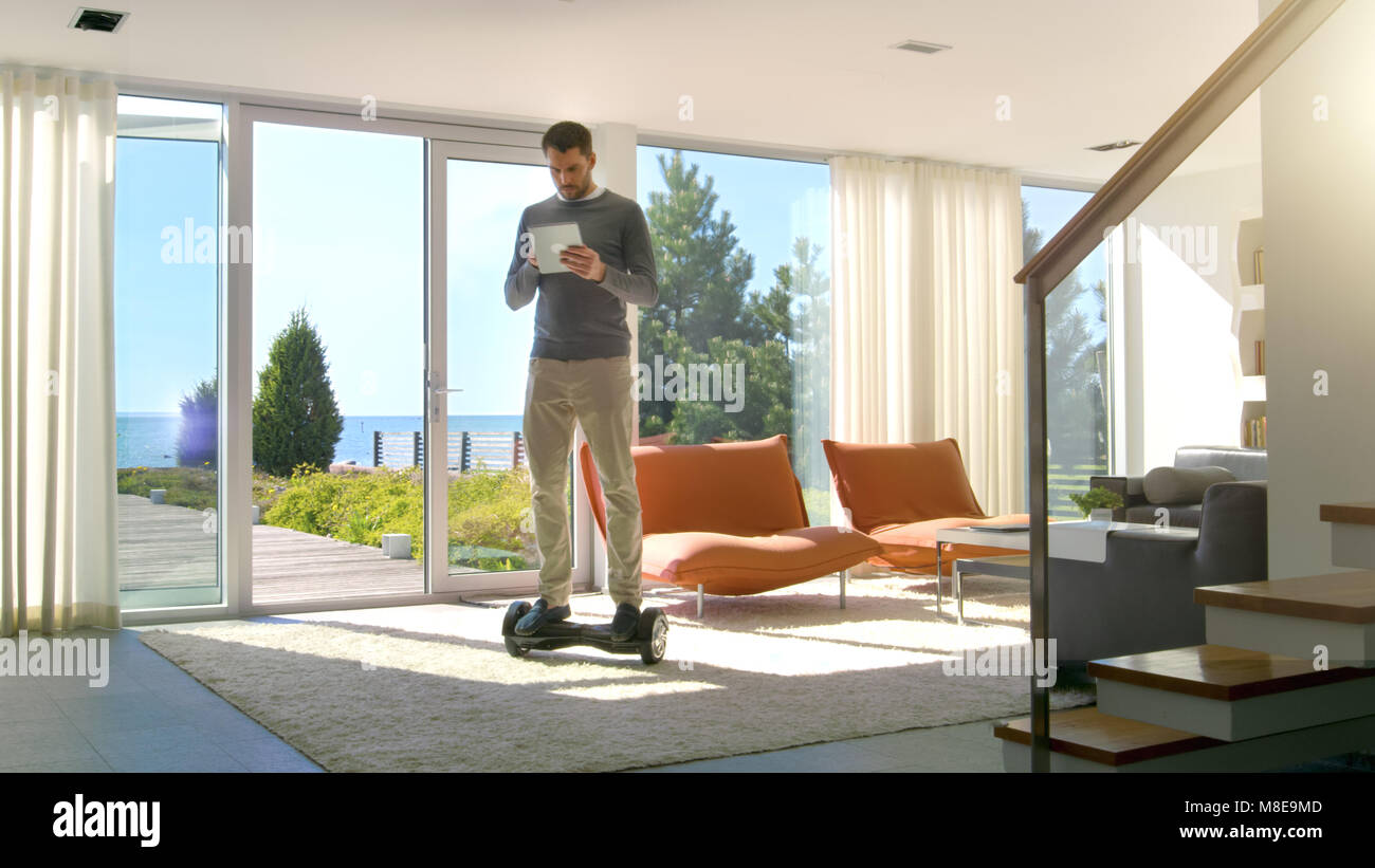 Tech Industry Businessman Working From Home Uses Tablet Computer and Rides Around the House on His Gyro Scooter. His Modern House has Seaside View. Stock Photo
