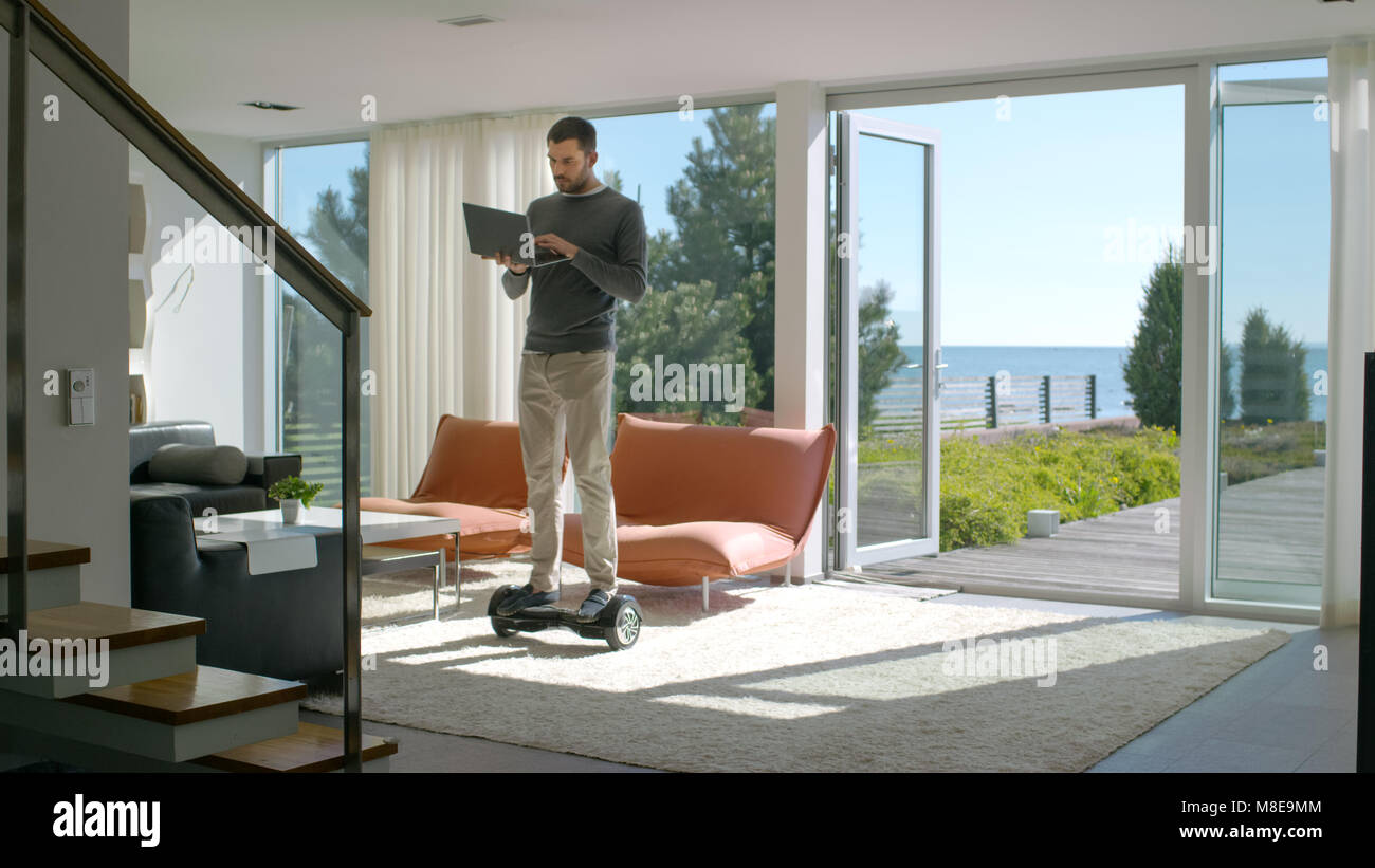 Tech Industry Businessman Working From Home Uses Laptop Computer and Rides Around the House on His Gyro Scooter. His Modern House has Seaside View. Stock Photo