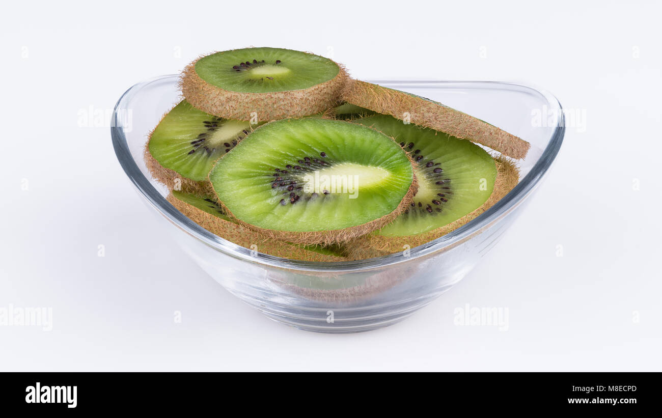 Green slices of kiwi in glass bowl. Fuzzy kiwifruit. Actinidia deliciosa. Close-up of heap of sliced tropical fruits with brown skin. White background. Stock Photo