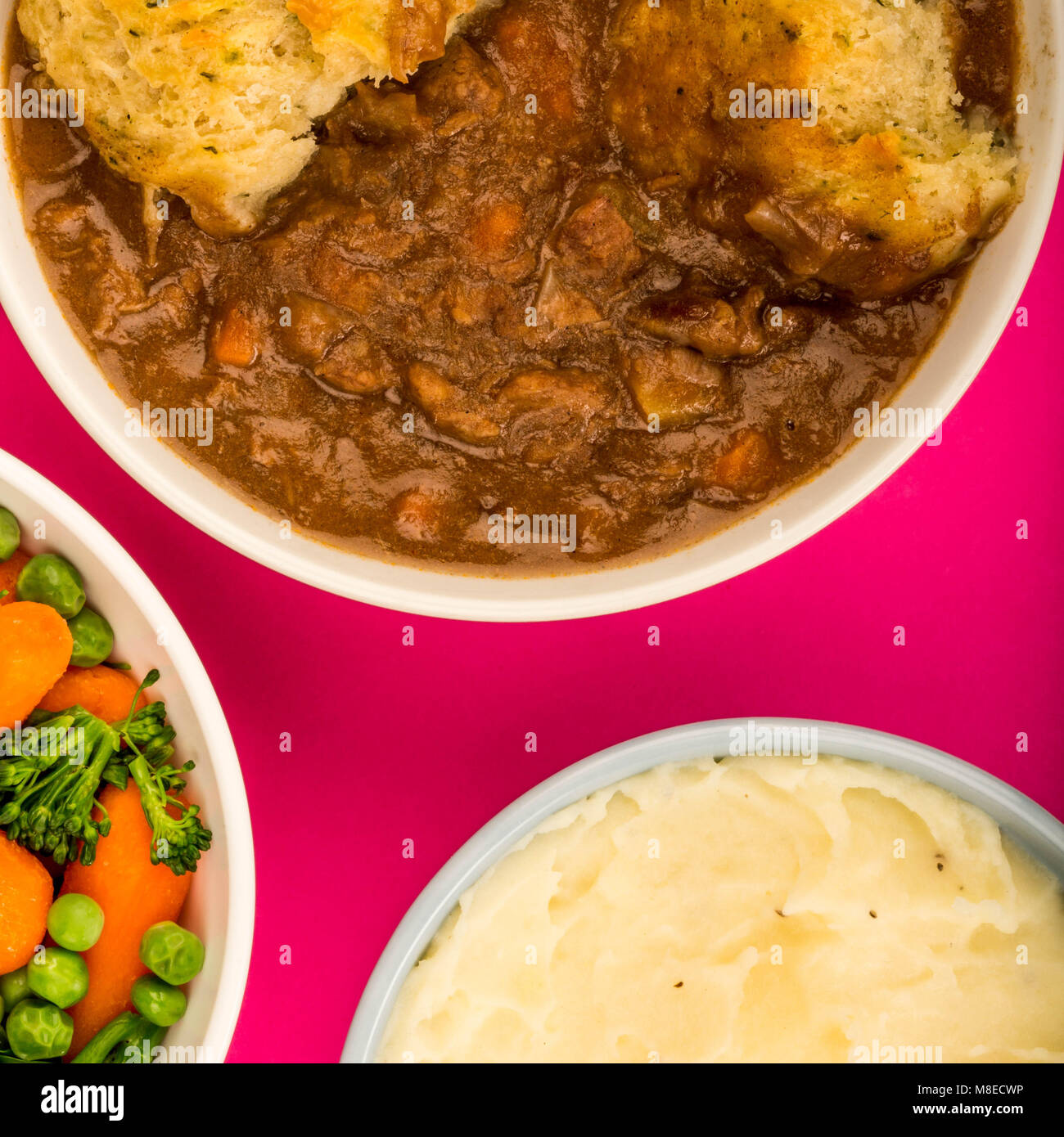 Traditional British Beef Casserole With Dumplings Against A Pink Background With A Bowl Of Mixed Vegetables And Mashed Potatoes Stock Photo