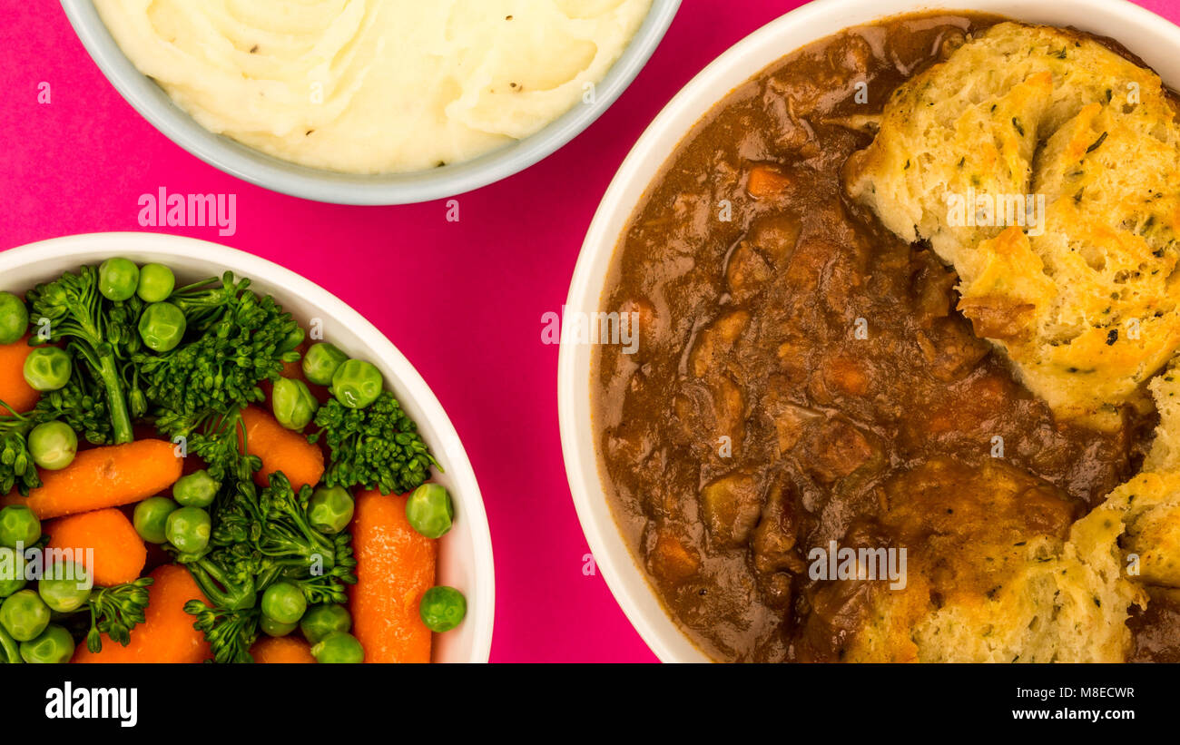 Traditional British Beef Casserole With Dumplings Against A Pink Background With A Bowl Of Mashed Potatoes And Mixed Vegetables Stock Photo