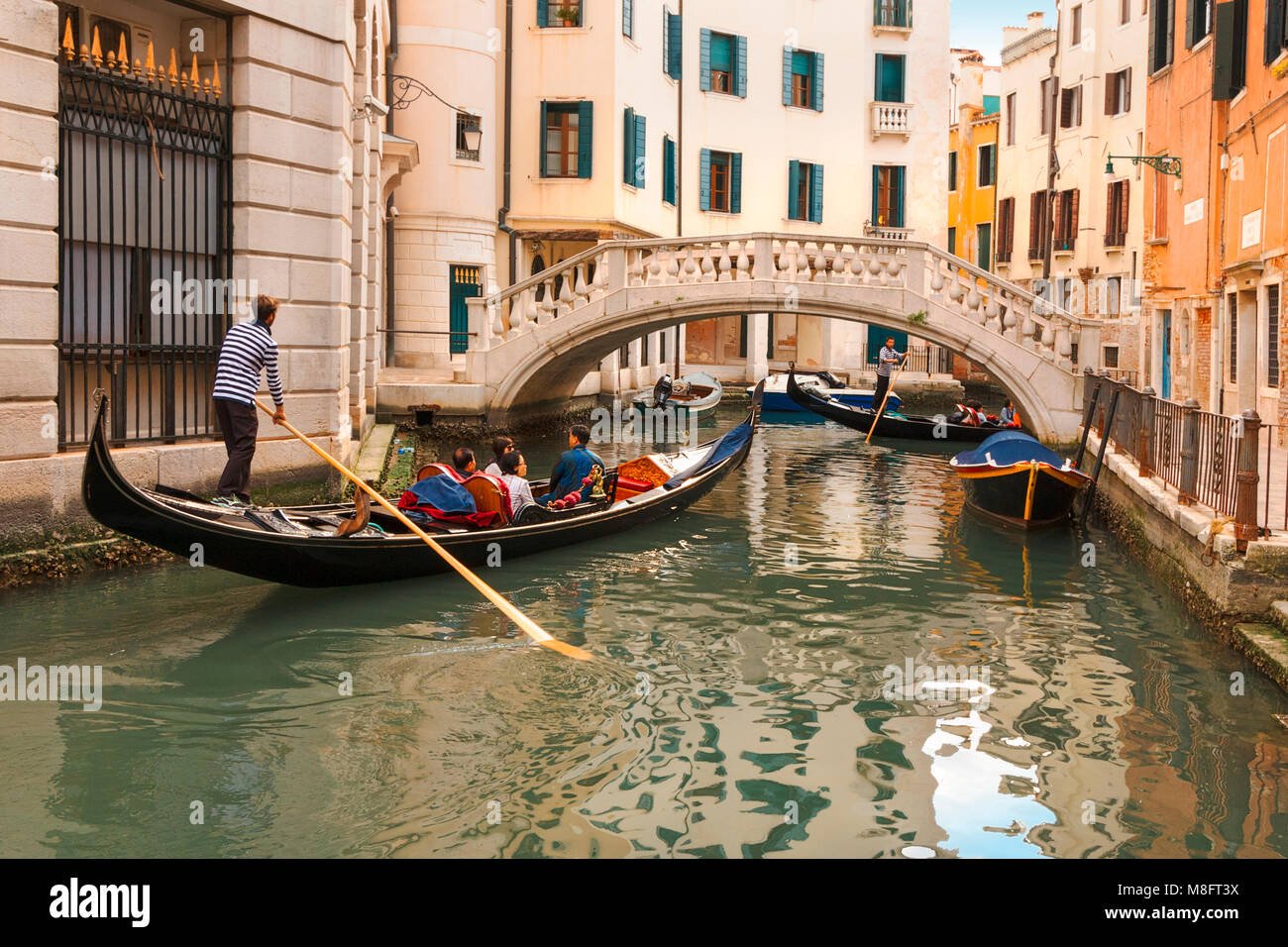 Colourful gondola journey with tourists and apartments with shutters overlooking an elegant white stone bridge spanning a narrow canal in Venice Stock Photo