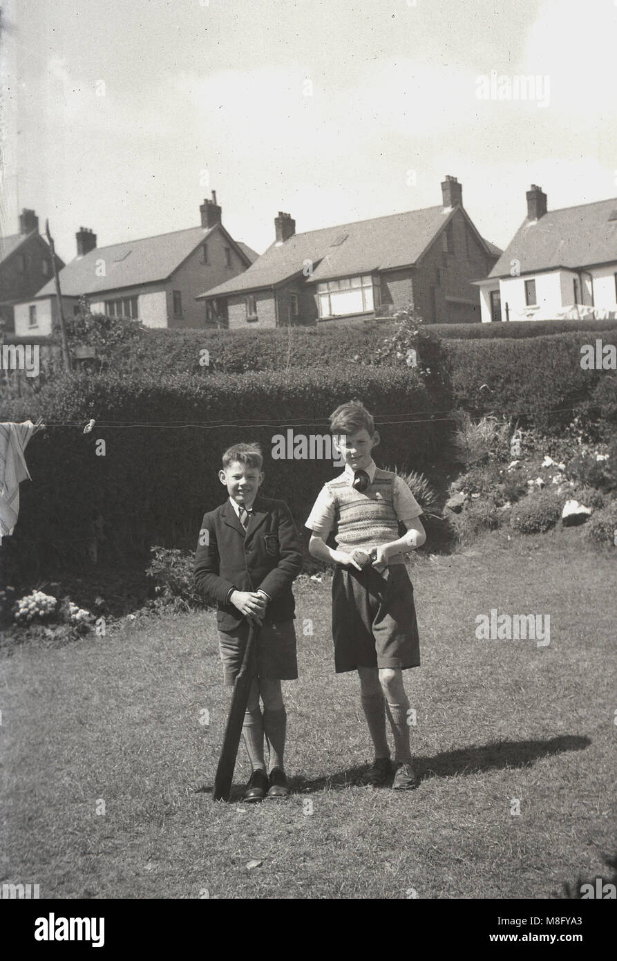 1950, historical, summertime and two smiling young boys, possibly brothers, standing together outside in a suburban back garden, the youngest holding an old cricket bat, England, UK. Stock Photo