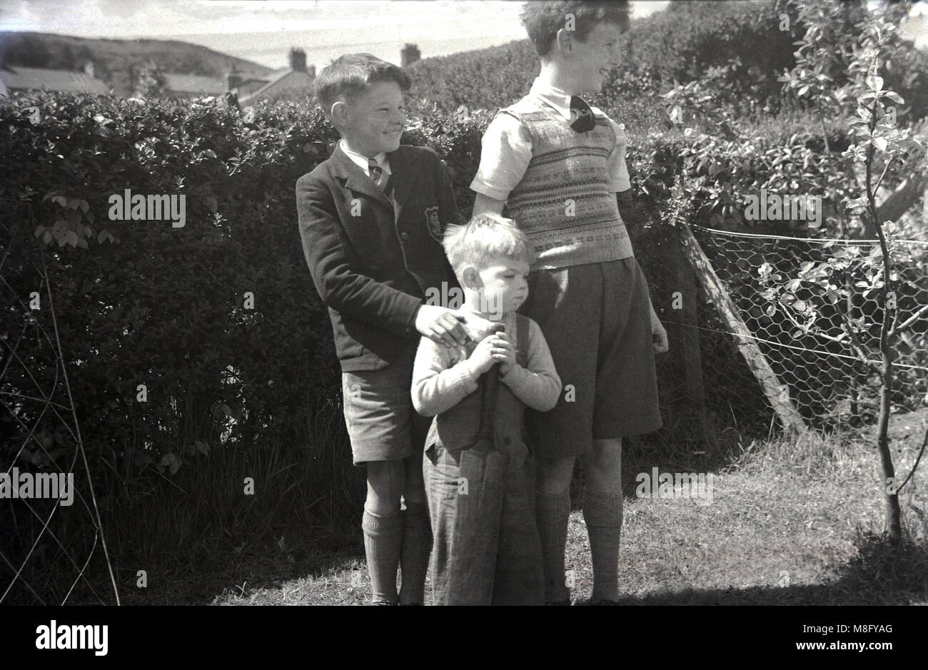 1950, historical, summertime and three happy, smiling young boys, possibly brothers, standing together outside in a back garden, the youngest holding an old cricket bat, England, UK. Stock Photo