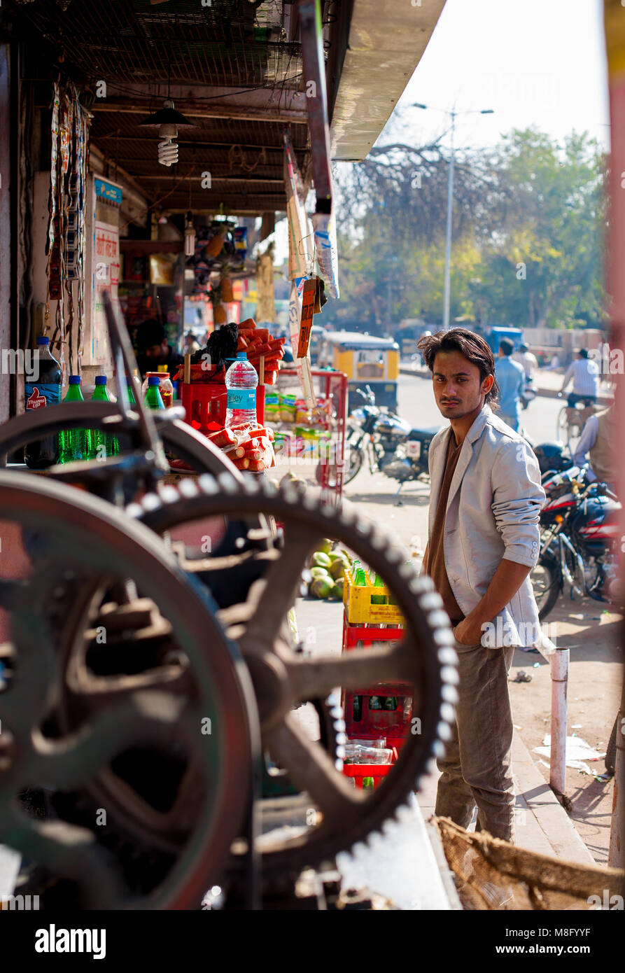 A young Indian man wearing light shirt with hands in his pockets, standing next to a sugarcane machine at his street shop on busy road in Jodhpur Indi Stock Photo