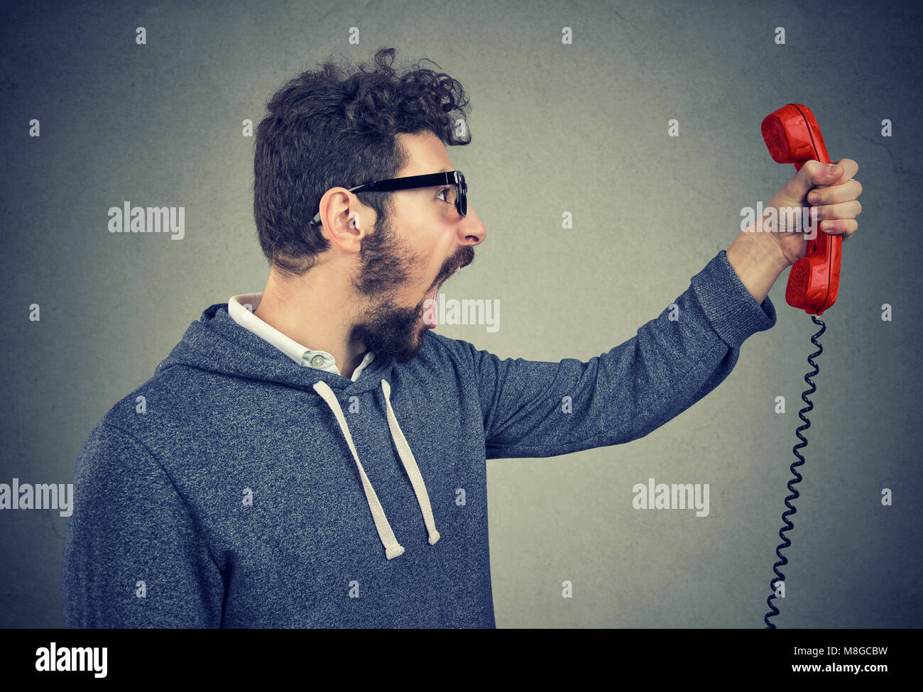 Side view of casual bearded man holding red telephone receiver and yelling in anger. Stock Photo