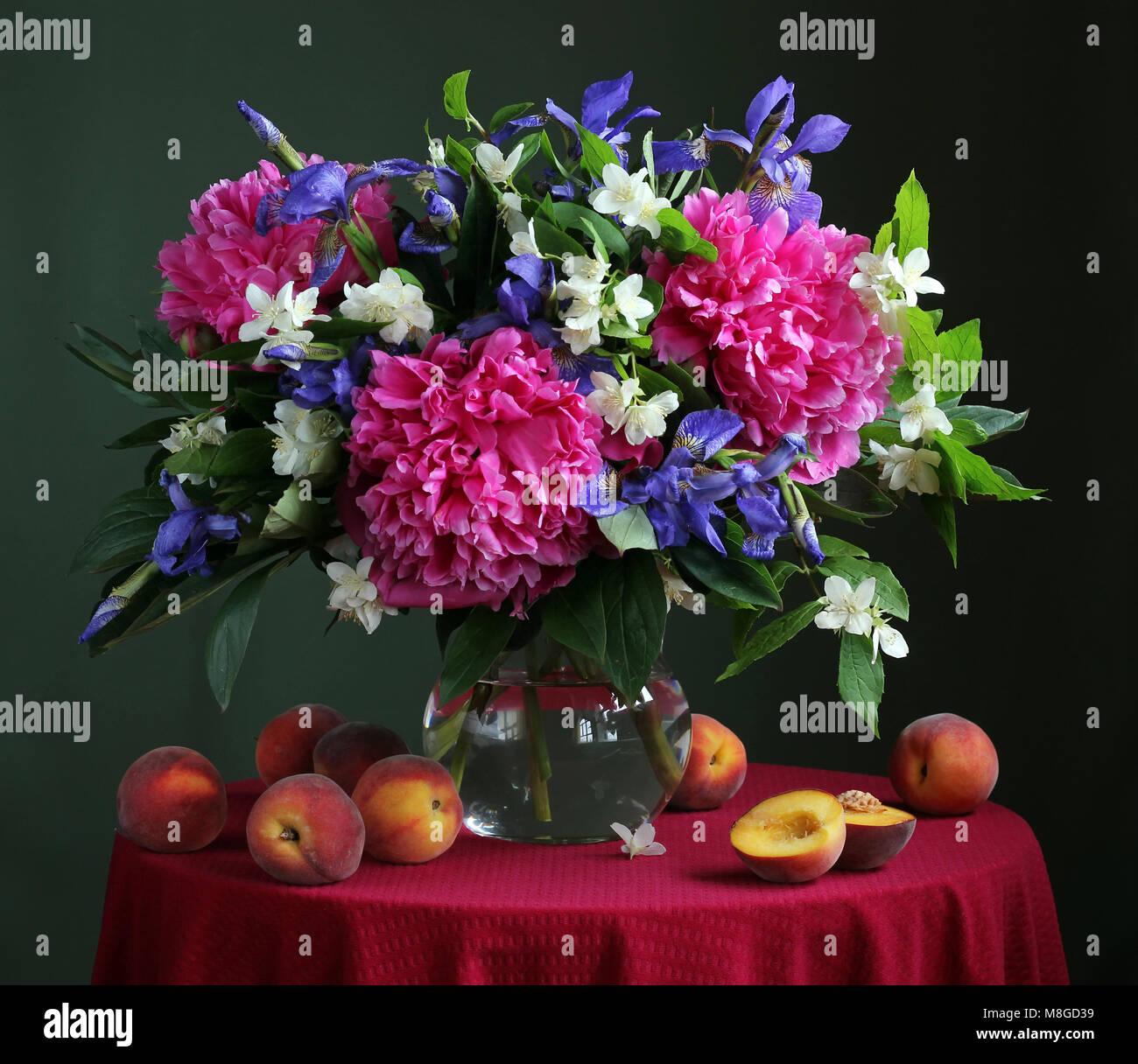 Bouquet of cultivated flowers in the jug and peaches on a round table with a red tablecloth. Still life with peonies, Jasmine and iris. Stock Photo