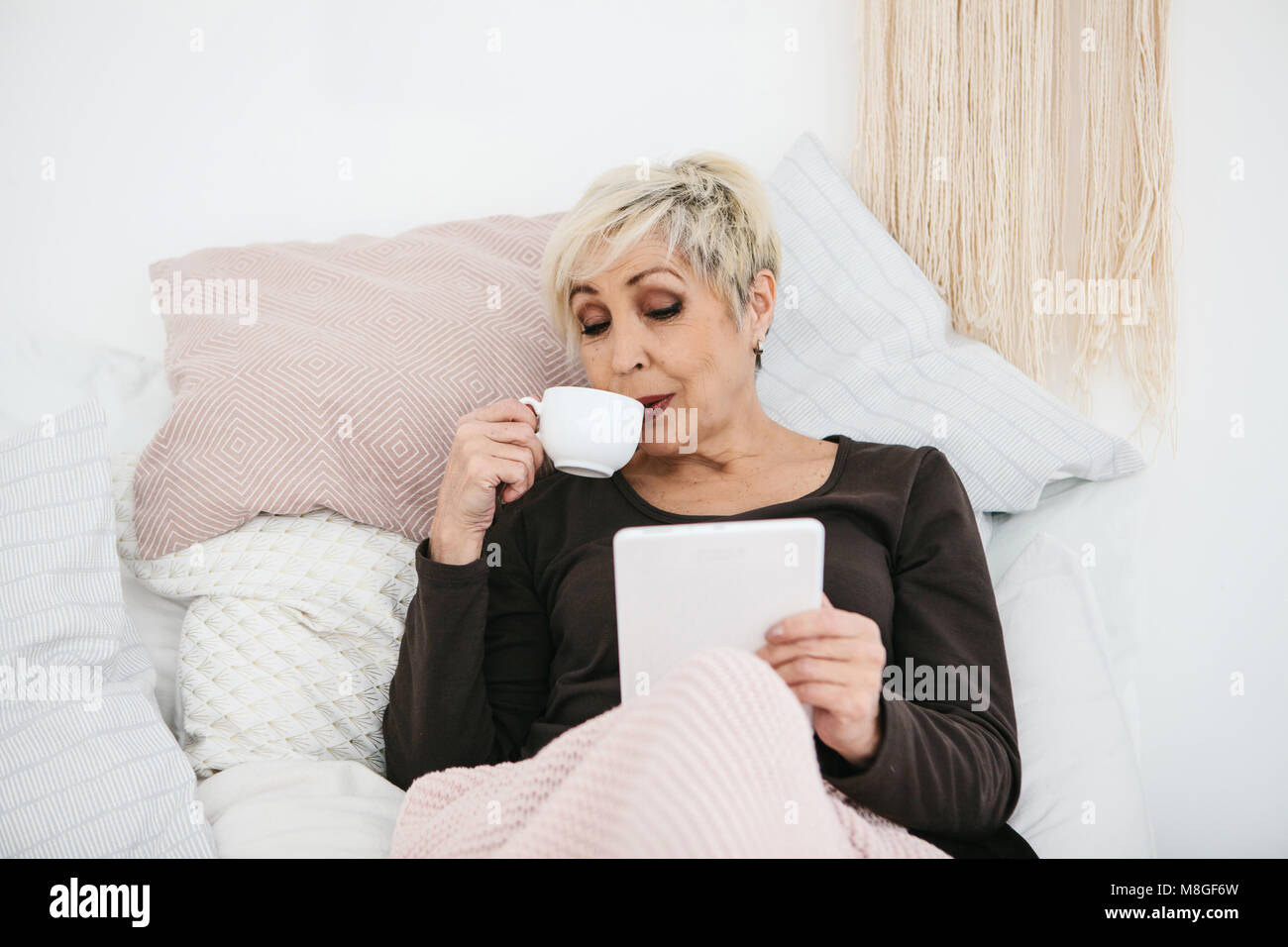 An elderly woman lying in bed drinking morning coffee uses a tablet to