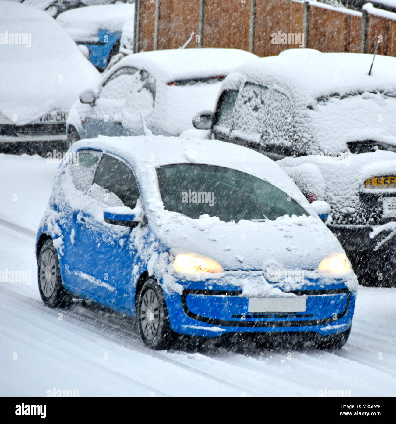 Travel scene blue car covered in snow headlights on driving in bad winter weather with more snowfall on snowy covered road Brentwood Essex England UK Stock Photo