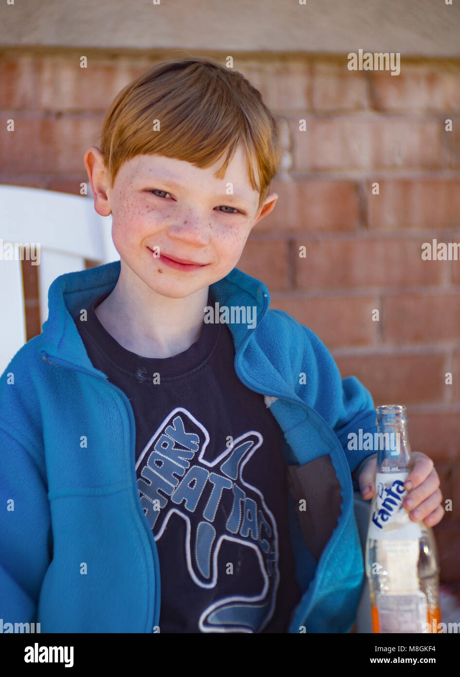 Boy with red hair and freckles, wearing a blue jacket, and holding a bottle of Orange Fanta soda. Stock Photo