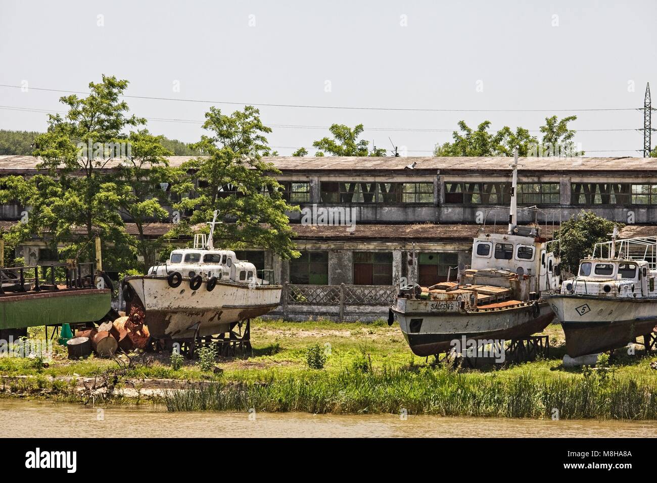 RUSTING SHIPS ON SHORE ABOVE THE DANUBE IN FRONT OF EMPTY AND ABANDONED CONCRETE BUILDING SULINA ROMANIA Stock Photo