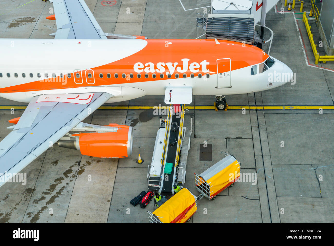 High perspective of baggage handlers picking up suitcases from a conveyor belt connected to an Airbus A320 easyJet airplane at London Gatwick's North Terminal Stock Photo