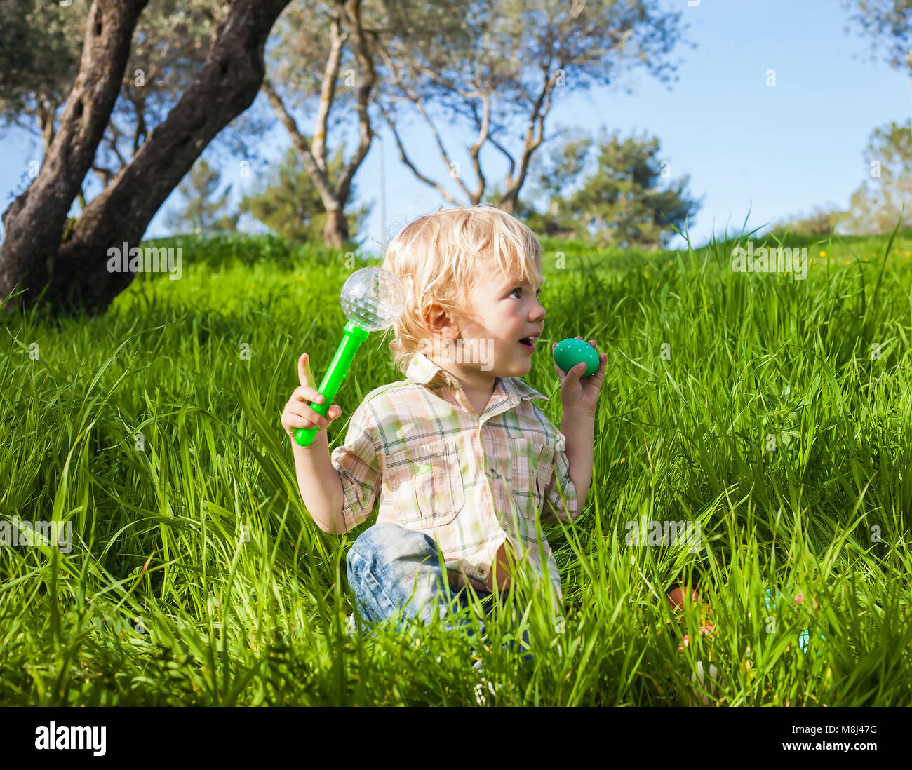 Funny toddler boy pretends to bite a colored Easter egg in the green grass Stock Photo