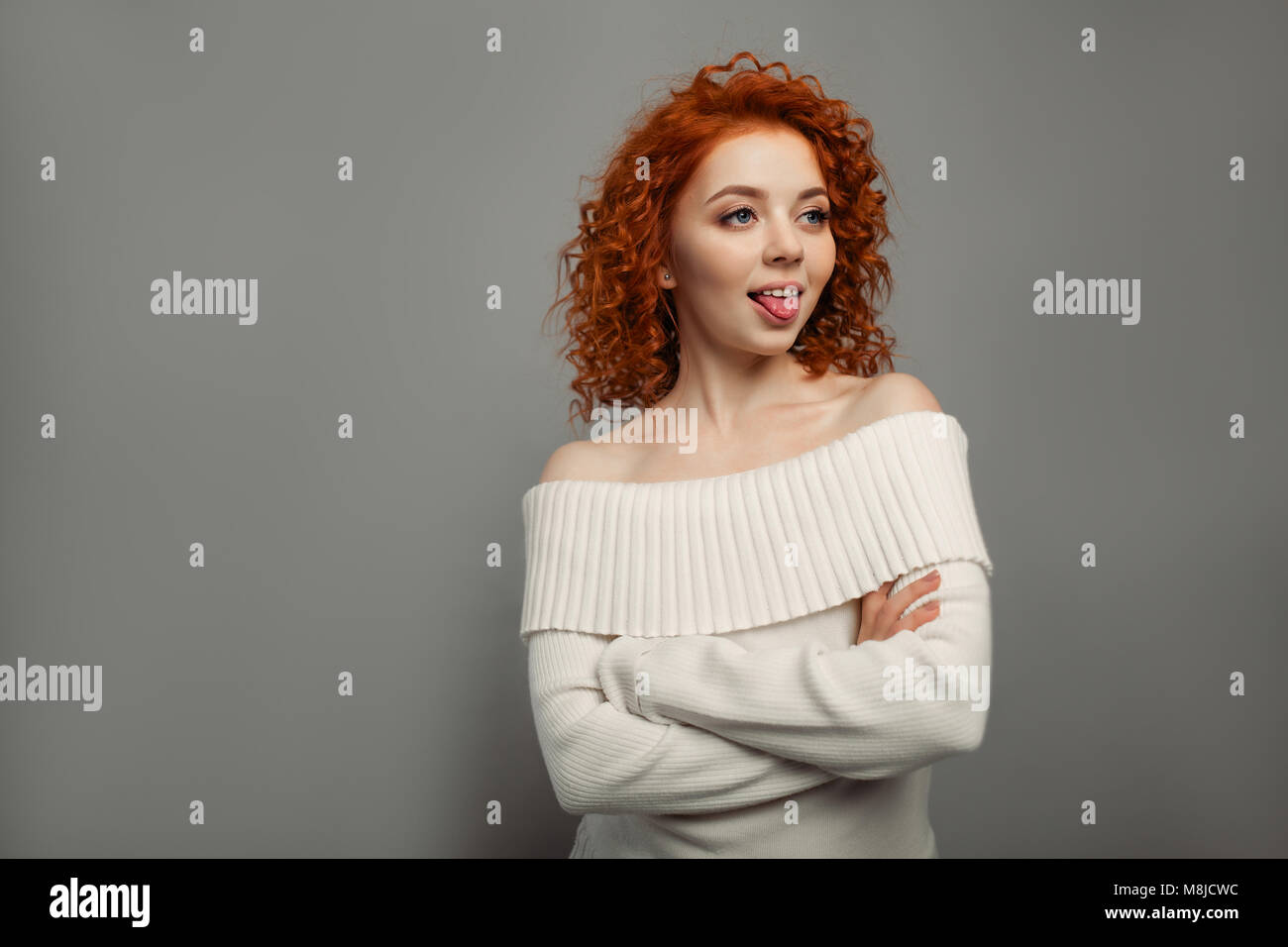 young cute redhead curly haired girl shows her tongue teasing on the gray backgroun in studio Stock Photo