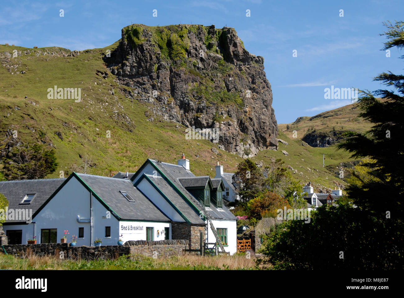 Bed & Breakfast establishment beneath large rocky outcrop on mountainside, Ellenabeich, Seil island, Scotland Stock Photo