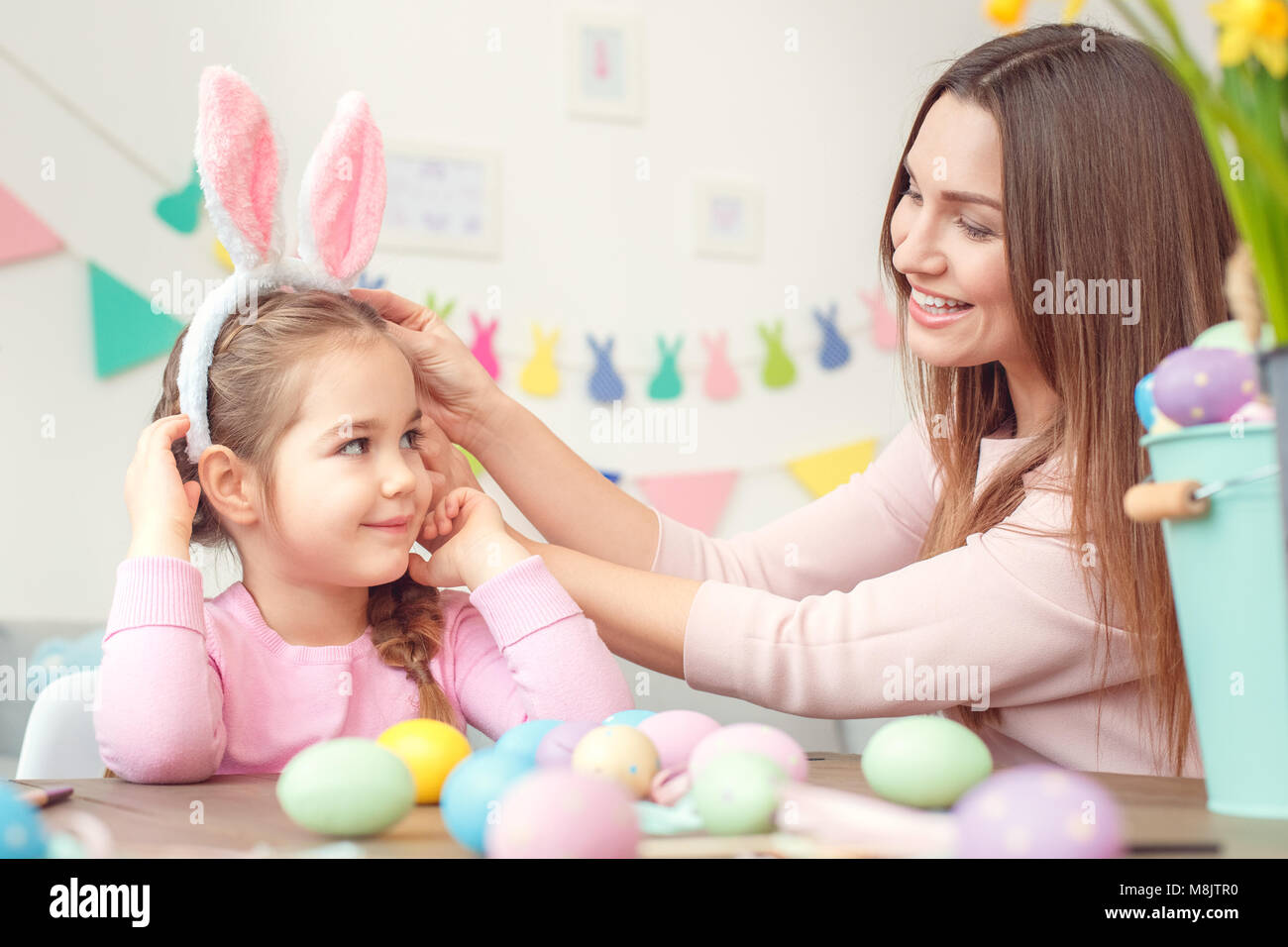 Young mother and little daughter wearing bunny ears easter celebration together at home sitting at the table mom touching bazel on girl's head smiling Stock Photo