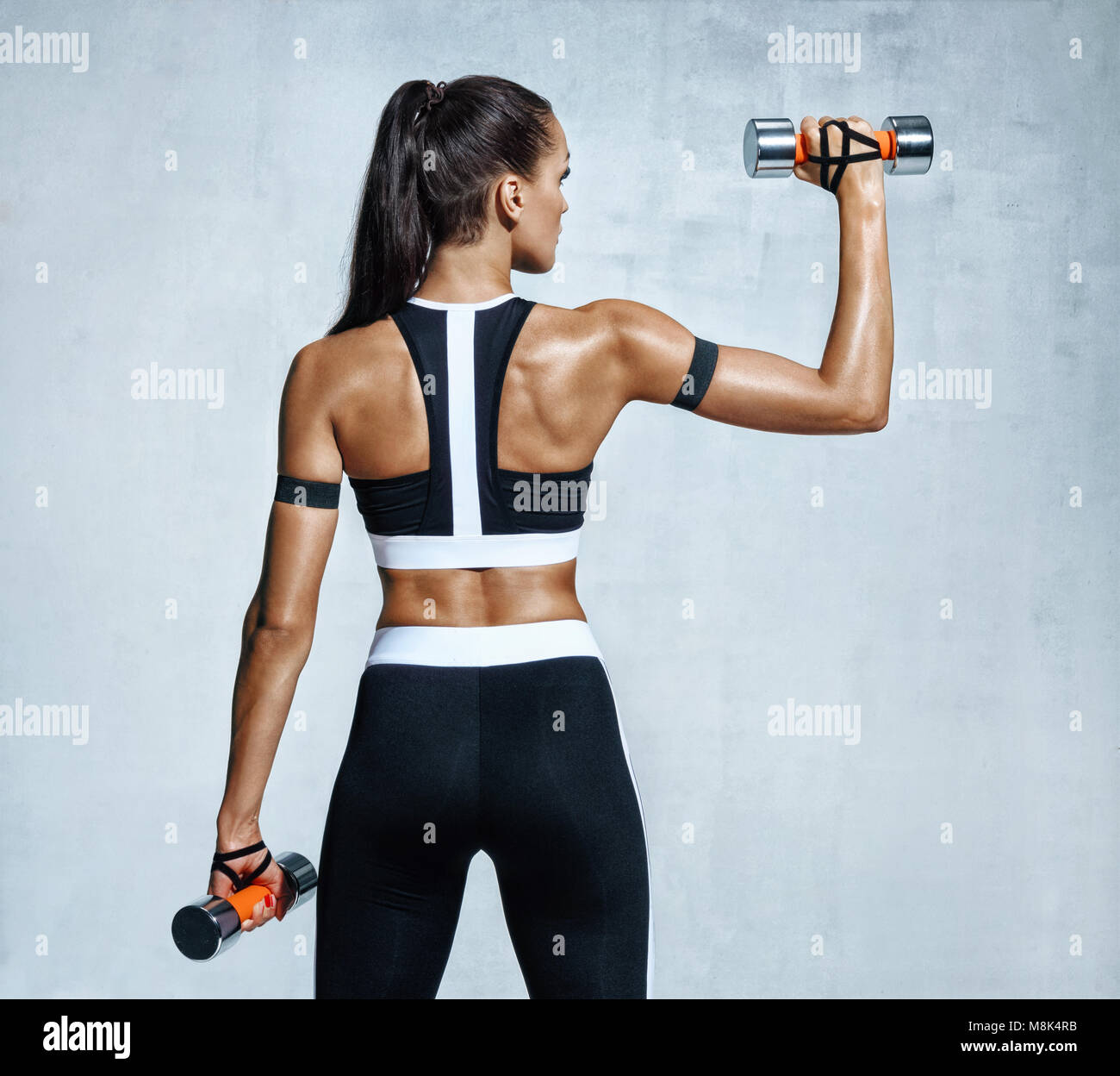 Strong woman working out with dumbbells, flexing her arm. Photo of sporty woman in sportswear on grey background. Rear view Stock Photo