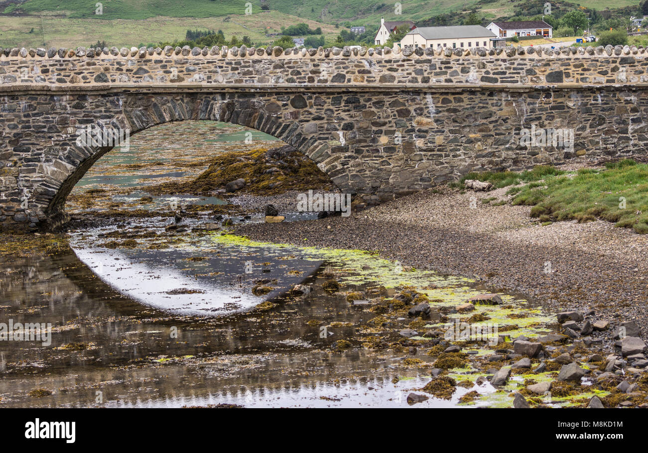 Dornie, Scotland - June 10, 2012: One bow of footbridge to Eilean Donan Castle. Half reflected on waters during low tide. Green hill with houses as ba Stock Photo