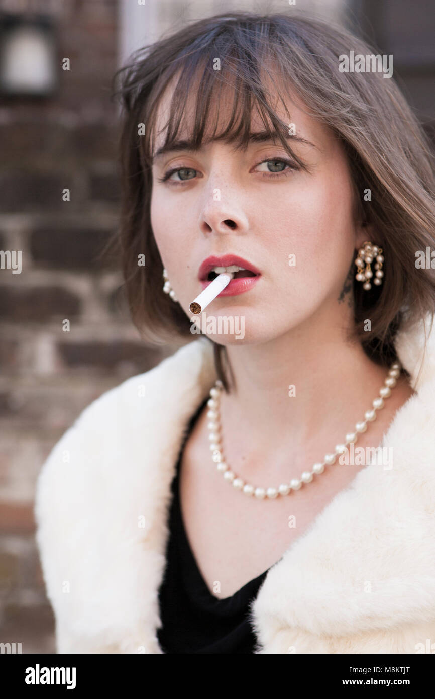 A well dressed woman wearing red lipstick and a pearl necklace, with an unlit cigarette in her lips. Stock Photo