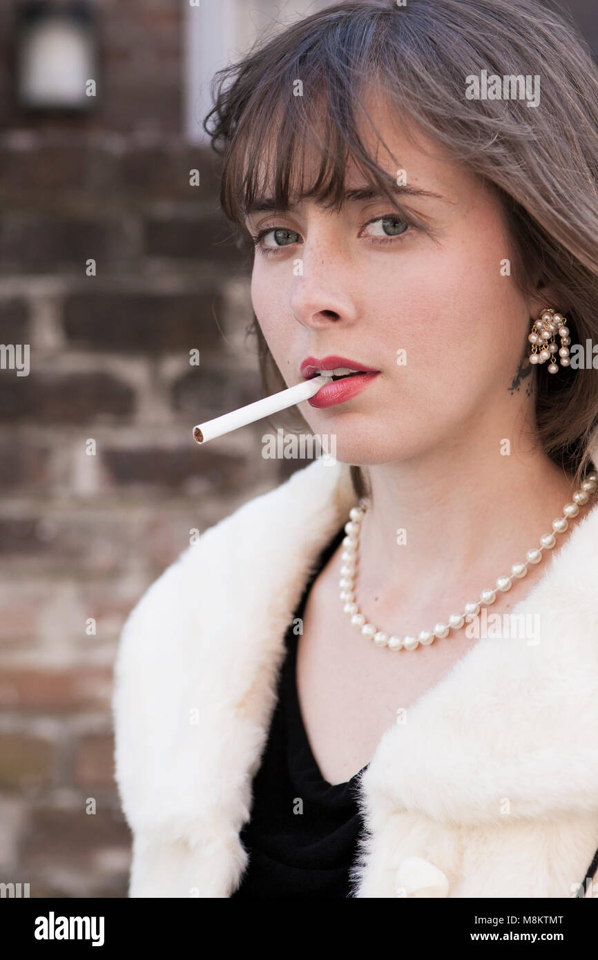 A well dressed woman wearing red lipstick and a pearl necklace, with an unlit cigarette in her lips. Stock Photo