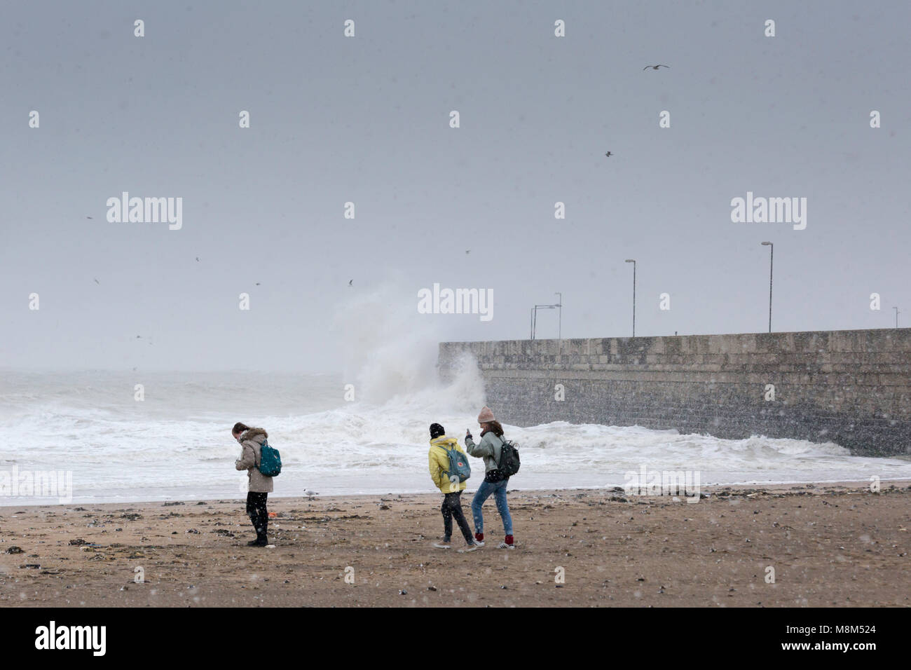 Ramsgate, Kent, UK 18th March, 2018. Three young women walk on Ramsgate beach during a snow storm while a large wave crashes into the harbour arm. Sue Holness Credit: Sue Holness/Alamy Live News Stock Photo