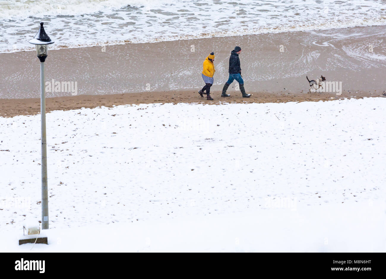 Couple walking with dog along seashore with Bournemouth beach seaside covered in snow at Bournemouth, Dorset, England UK in March Stock Photo