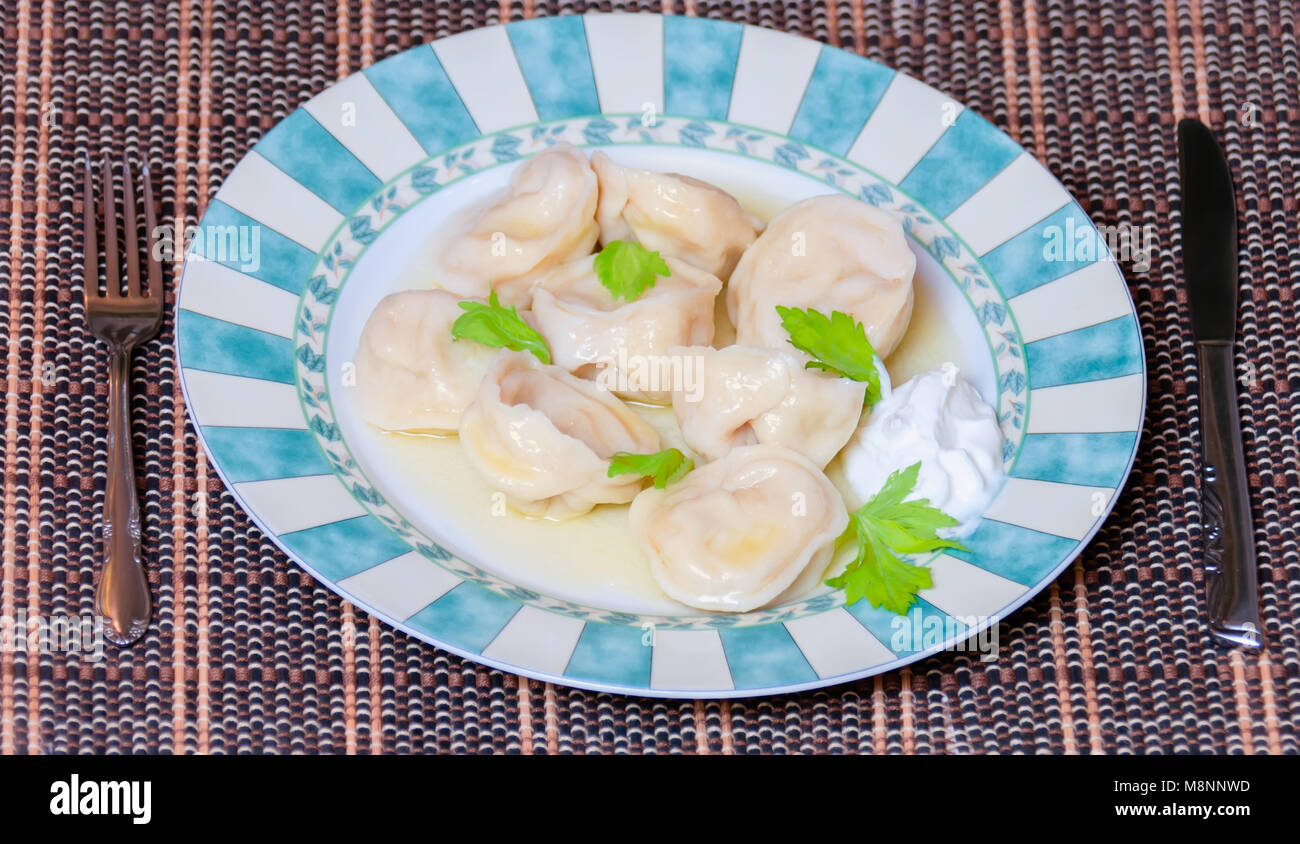 appetizing boiled dumplings with sour cream on a large beautiful plate with a green fresh leaf, a metal knife and fork lie on the edges, a rattan mat  Stock Photo