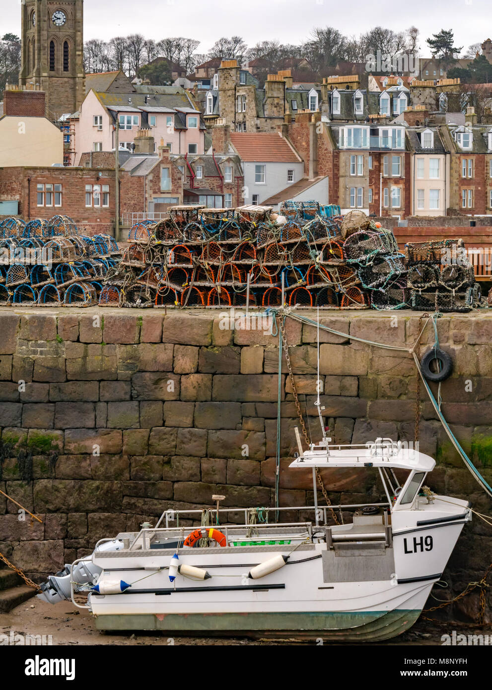 North Berwick, East Lothian, Scotland, United Kingdom,  Boat stranded in harbour at low tide with lobster creels on pier. Seaside houses in background Stock Photo
