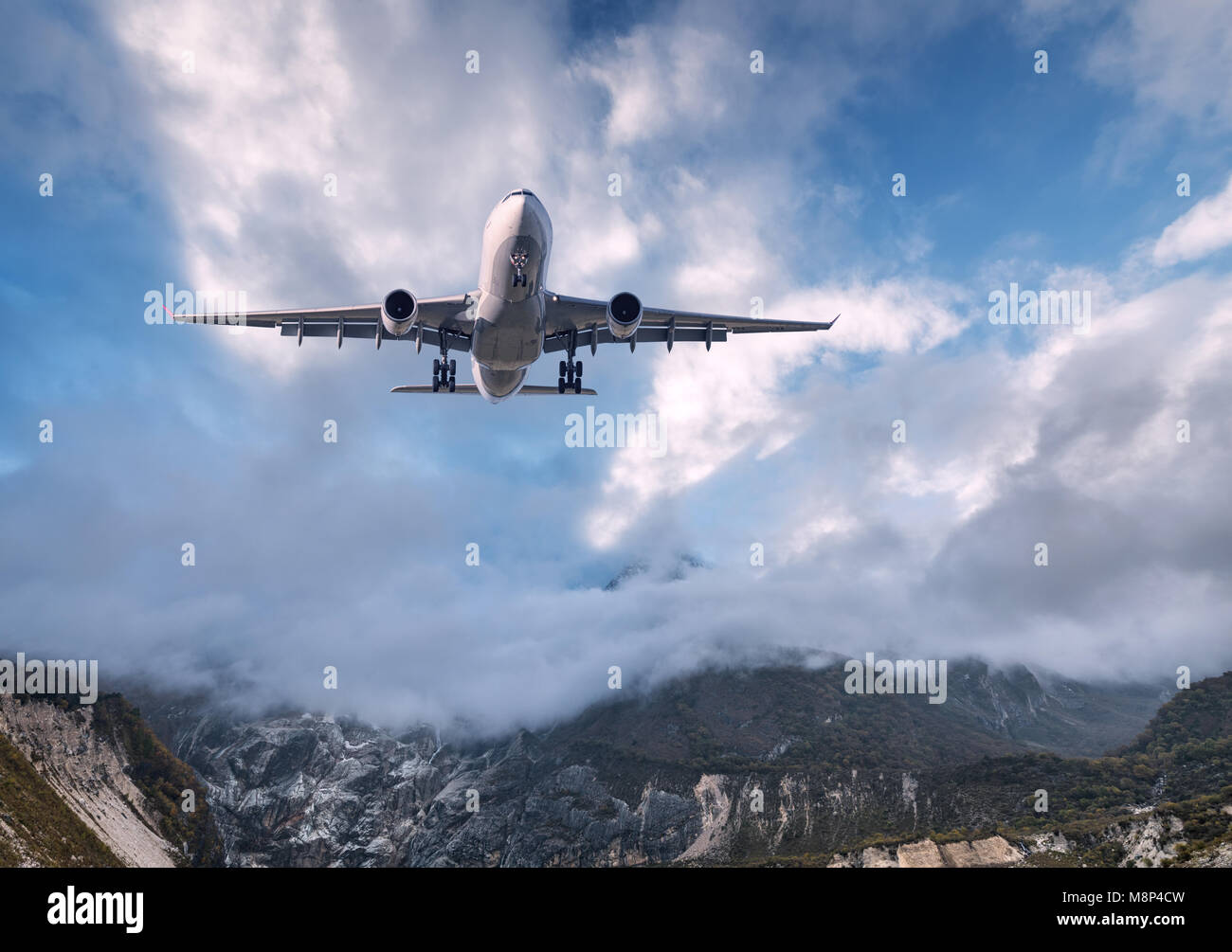 Big white airplane is flying over rocks at sunset. Landscape with passenger airplane, mountains, colorful blue sky with clouds. Passenger aircraft is  Stock Photo