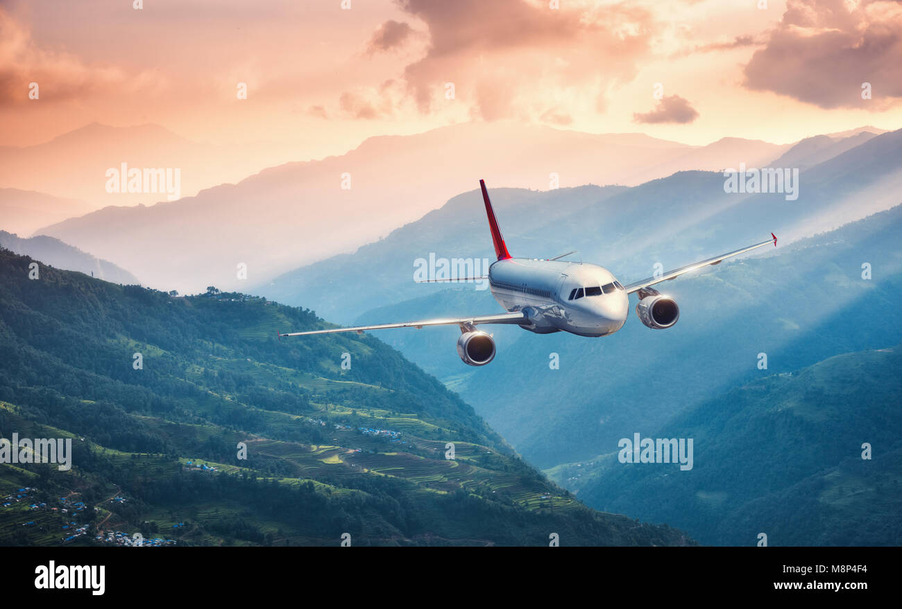 Aircraft is flying over green hills against mountains with yellow sunbeams at sunset. Landscape with passenger airplane, colorful sky, village. Passen Stock Photo