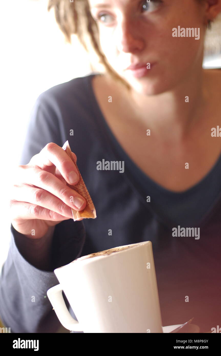Woman in in a blue top in a cafe emptying a sachet of sugar into a white cup of coffee while glancing out of the window Stock Photo